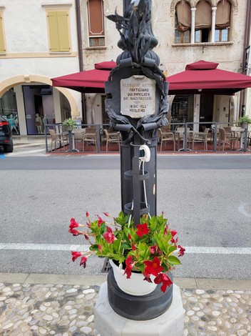 Targa commemorativa dedicata a Pellegrini, con decorazioni e fiori, in un contesto urbano.