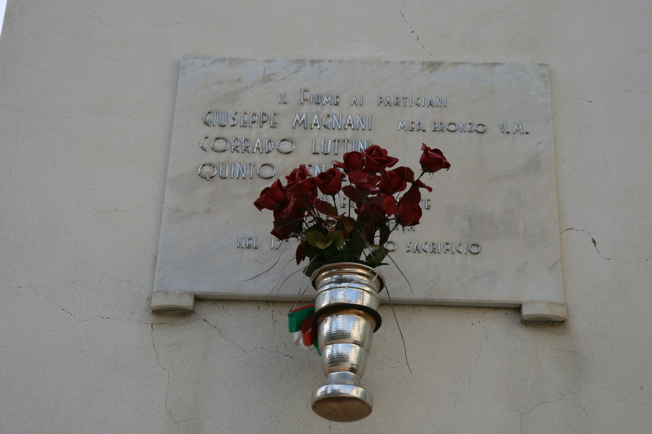 Lapide commemorativa dedicata a Giuseppe Magnani, Corrado Luttini e Quinto Genalti, con fiori rossi.