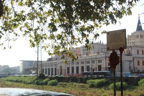 Lapide commemorativa in un contesto urbano, con edifici e vegetazione circostante visibili sullo sfondo.