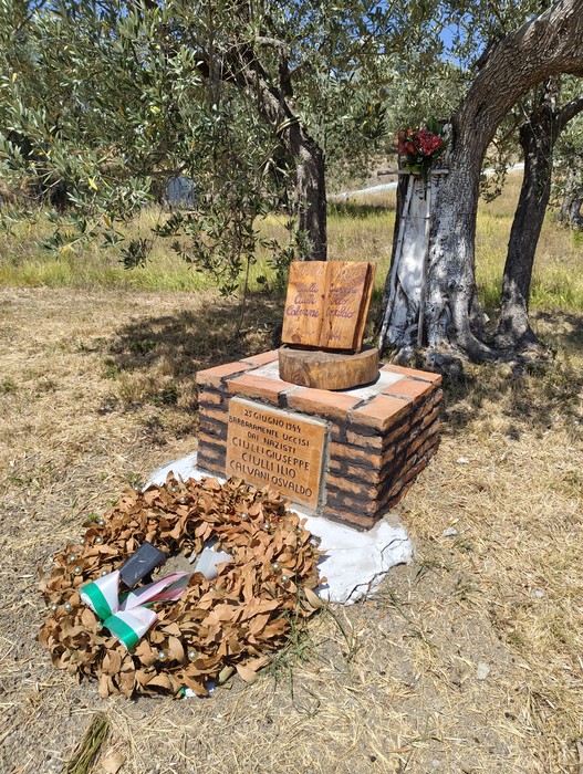 Cippo in mattoni con lapide incisa, corona di alloro e tavoletta di legno, sotto alberi d'ulivo.