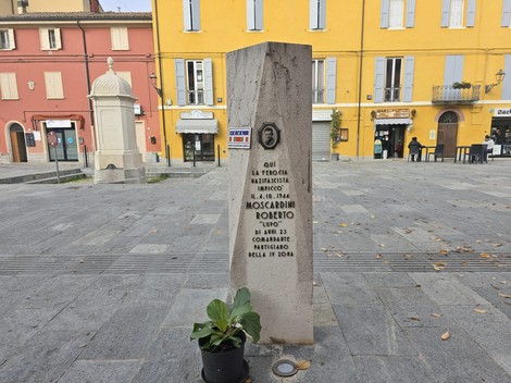Cippo commemorativo dedicato a Moscardini, situato in una piazza con edifici colorati sullo sfondo.
