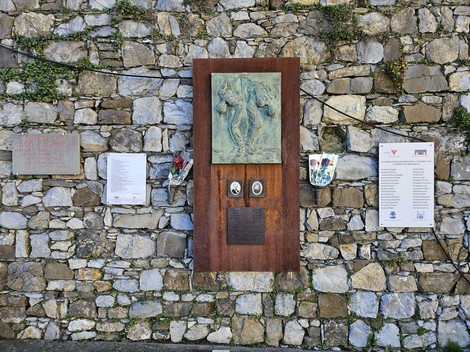 Targhe informativa e artistica su un muro di pietra a Piazza del Popolo, con elementi decorativi e varie iscrizioni.