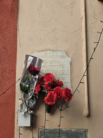 Lapide commemorativa con fiori rossi, affissa su un muro, in memoria di un patriota.