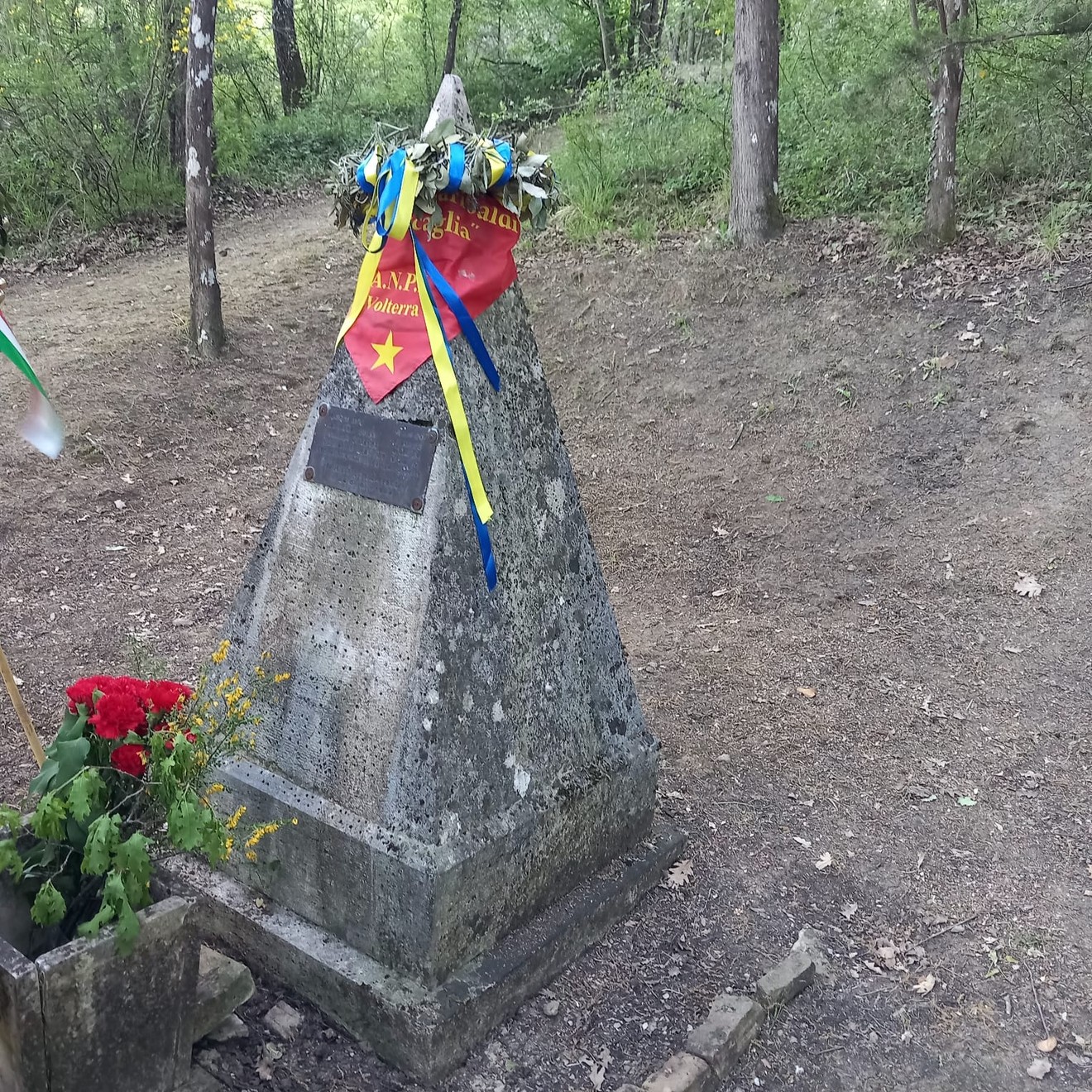 Cippo commemorativo circondato da vegetazione, decorato con nastri e fiori in ricordo di un evento storico.