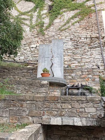 Monumento ai caduti con una stele in pietra affiancata da una pianta, situato su un muro di pietra.