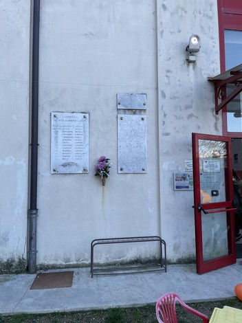 Lapidi commemorative ai caduti di Savonera, con fiori in memoria dei defunti, su una parete esterna.