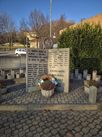 Monumento in pietra con lapidi incise e corona di fiori, in un'area pavimentata circondata da siepi.