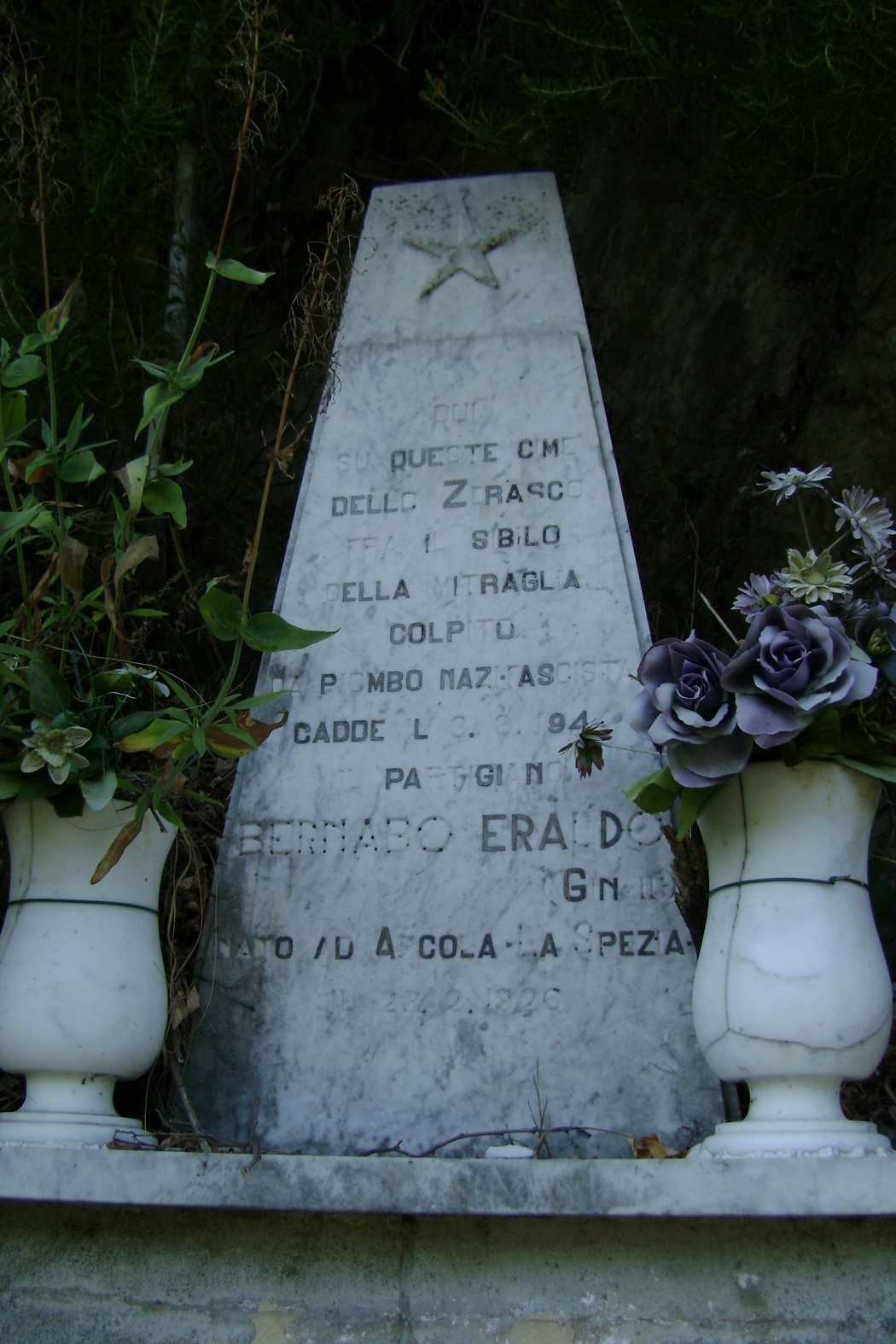 Cippo commemorativo dedicato a Bernabo, circondato da fiori e vegetazione, simbolo di memoria storica.
