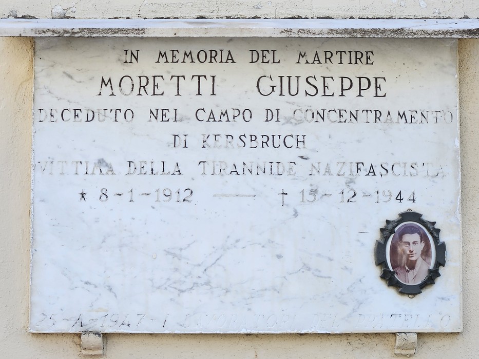 Lapide in marmo su parete esterna, con iscrizione e medaglione fotografico ovale di un uomo.