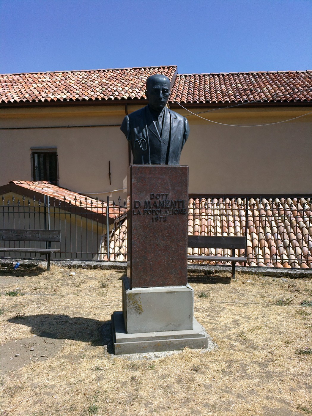 Monumento a Manenti: busto commemorativo con base in pietra, immerso in un paesaggio sereno.