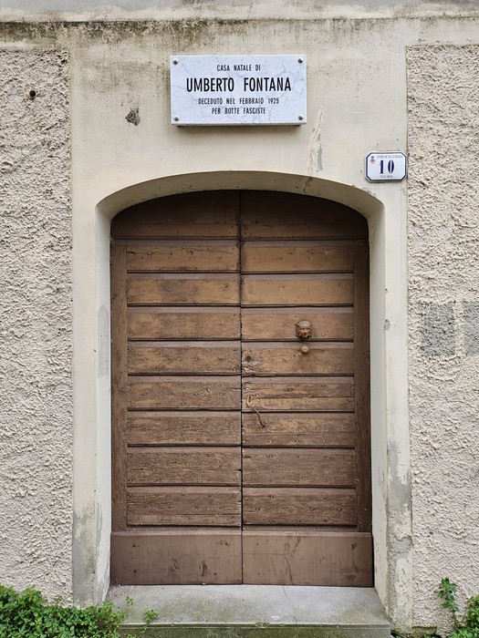 Ingresso con porta in legno e targa commemorativa di Umberto Fontana su una parete intonacata.