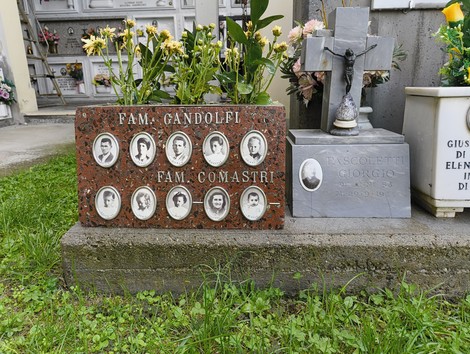 Monumento commemorativo con nomi di caduti, circondato da fiori in un cimitero. Rappresenta il ricordo dei defunti.