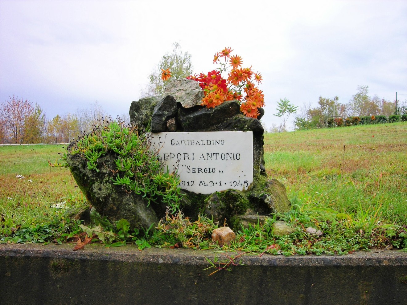 Cippo commemorativo dedicato a Lepori Antonio, decorato con fiori, immerso in un prato verde.