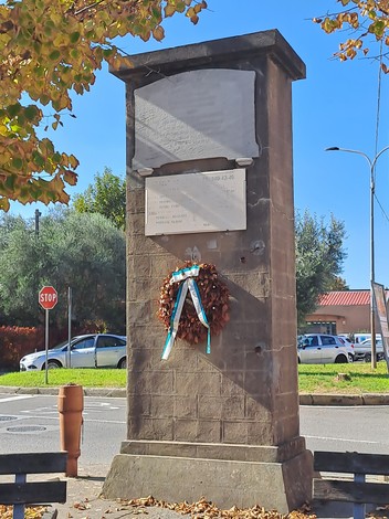 Lapide commemorativa ai caduti di Mezzana, adornata con una corona d'alloro, in un contesto urbano.