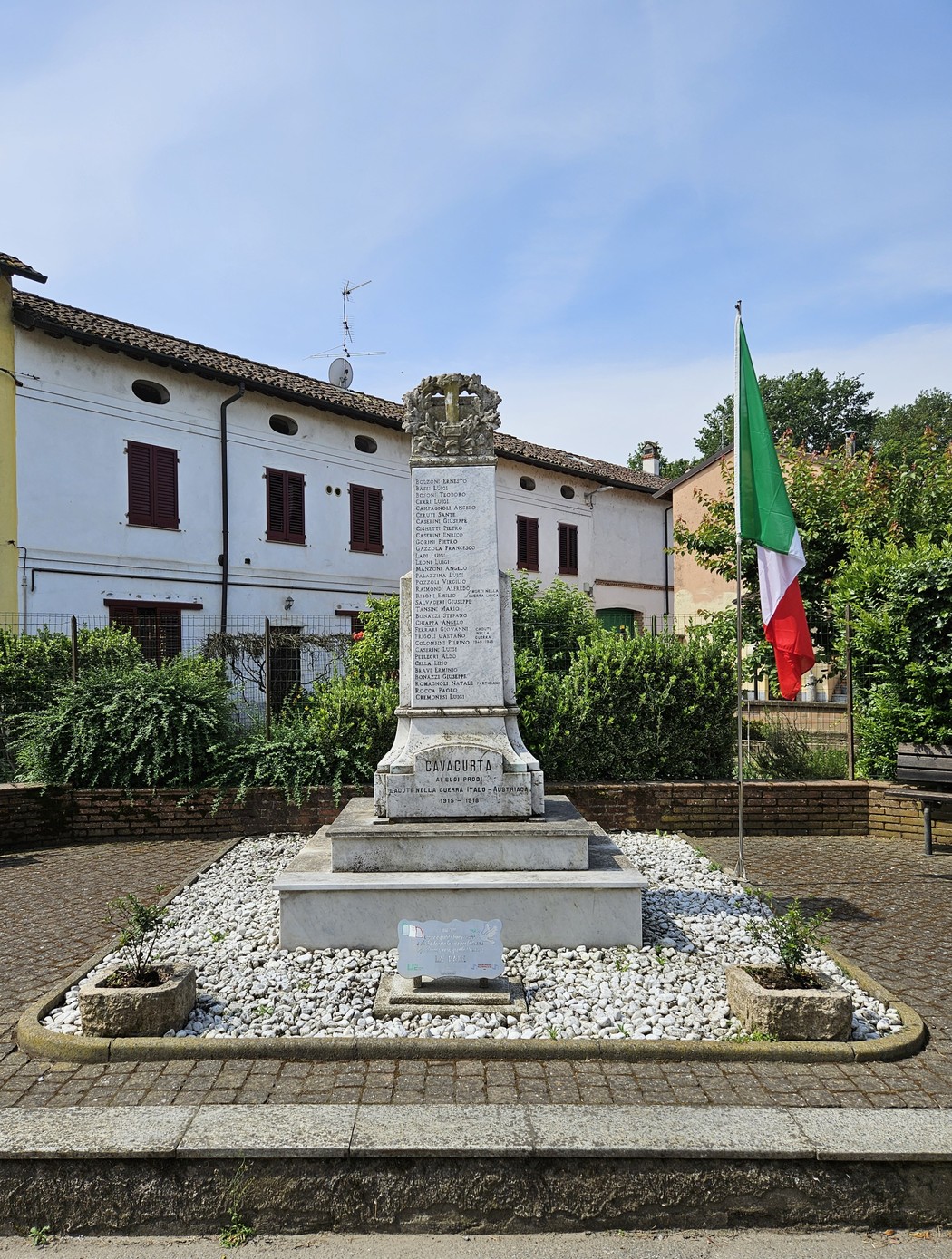 Monumento ai Caduti di Cavacurta, con bandiera italiana e circondato da aiuole e un pavimento di ciottoli.