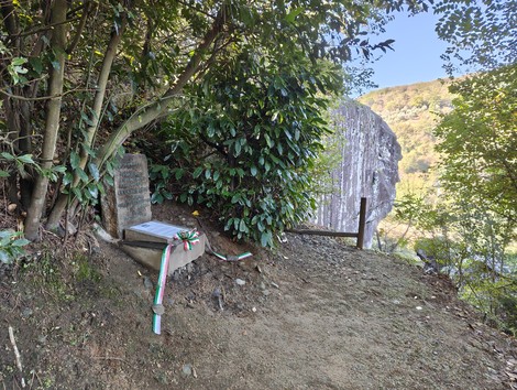 Cippo commemorativo in un'area verde, circondato da alberi e vicino a una roccia imponente.