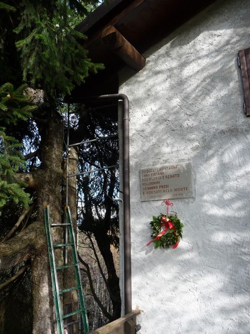 Lapide commemorativa con bronzo e una corona di fiori, apposta su un muro vicino a un albero.