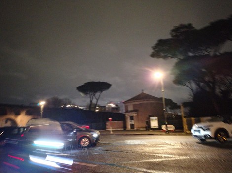 Cimitero di guerra del Commonwealth in una serata buia, con automobili in movimento e alberi sullo sfondo.