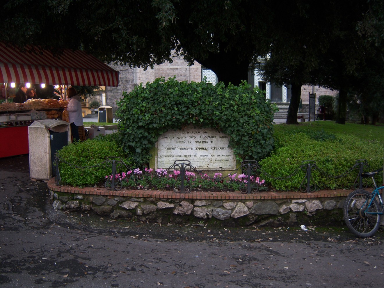 Monumento dedicato a Baronti e Spinelli, incorniciato da piante e fiori in un'area verde.