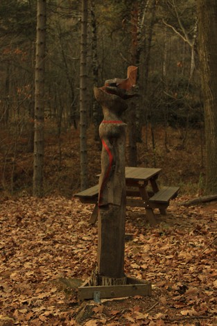 Scultura in legno a Gabarini, immersa in un ambiente boschivo con foglie secche e panchina in legno.