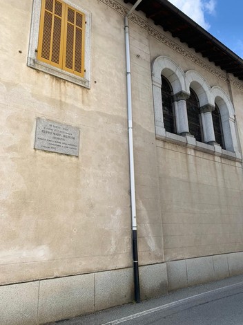 Lapide commemorativa su un muro esterno con finestre architettoniche, in un contesto urbano.