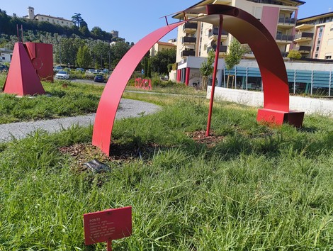 Scultura rossa su prato, con forme geometriche che interagiscono con l'ambiente circostante, evocando resistenza e collettività.