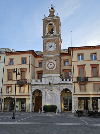 Lapide in memoria dei caduti, situata in una piazza storica con edifici colorati e un'orologio elegante.