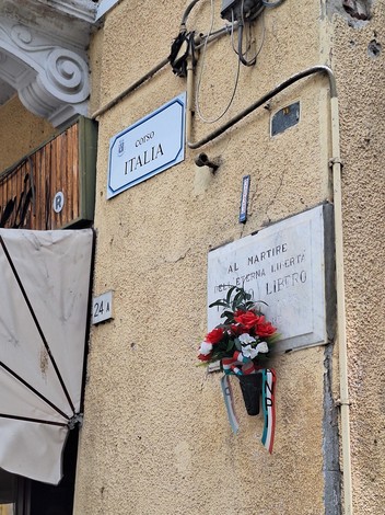 Lapide commemorativa su una parete di un edificio, con fiori e decorazione tricolore.
