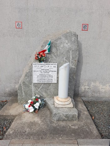 Lapide commemorativa con fiori e bandiera, in memoria di Boetto e Boero, in un contesto urbano.