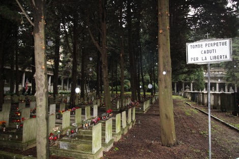 Campo di tombe con fiori e una segnaletica che indica "Tombe perpetue dei caduti per la Libertà" tra alberi.