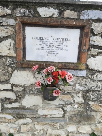 Lapide commemorativa di Ghiandelli, con fiori decorativi, affissa su una parete di pietra.