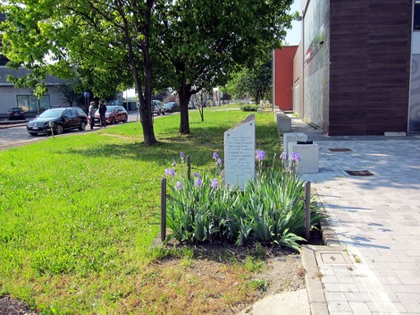 Cippo commemorativo circondato da fiori in un'area verde, con alberi e un marciapiede sullo sfondo.