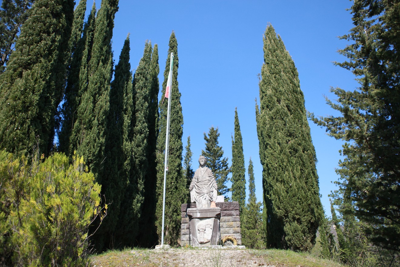 Monumento ai caduti di Purocielo circondato da alberi di cipresso e con una bandiera tricolore in evidenza.
