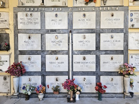 Parete con sedici lapidi marmoree, ciascuna con fototessera e iscrizione, scritta "Caduti per la Libertà" e fiori sottostanti.