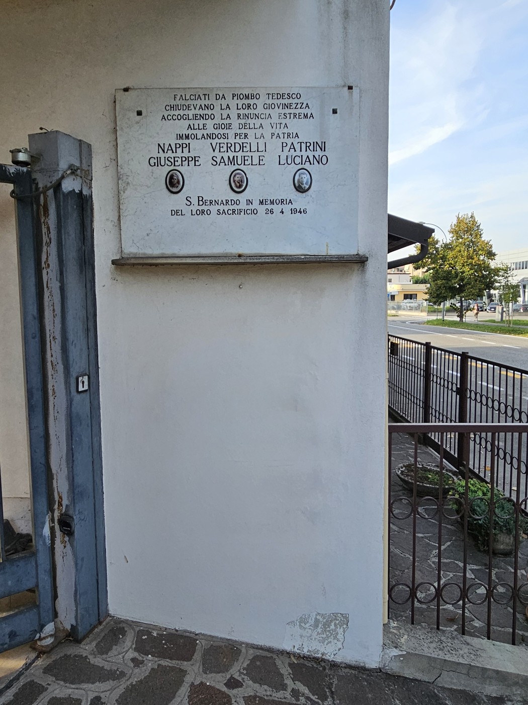 Lapide bianca con iscrizione e tre fototessere, murata su una parete esterna accanto a un cancello in ferro.