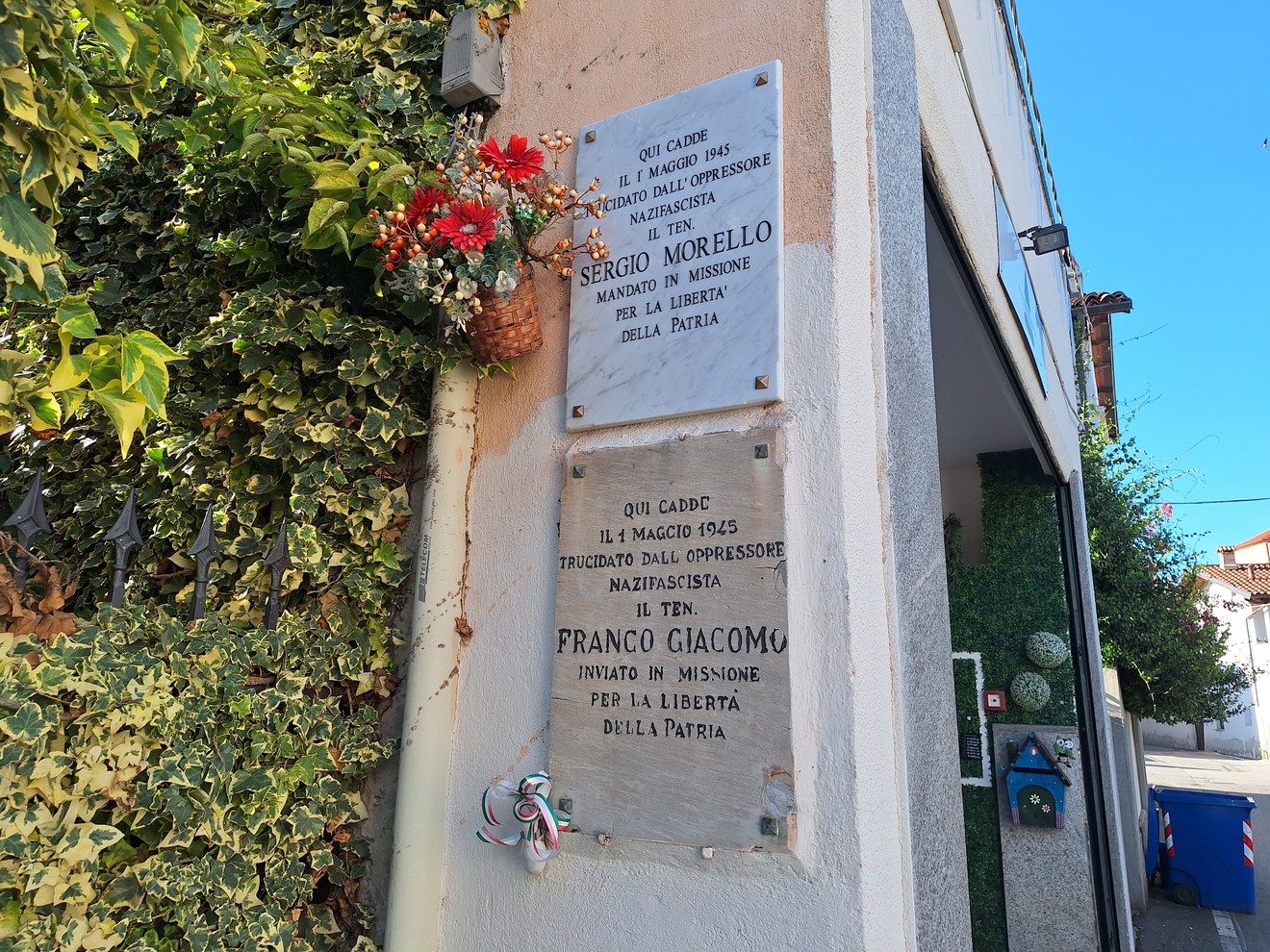 Due lapidi in marmo sulla parete d'angolo di un edificio, con fiori e fascia tricolore sotto la targa inferiore.