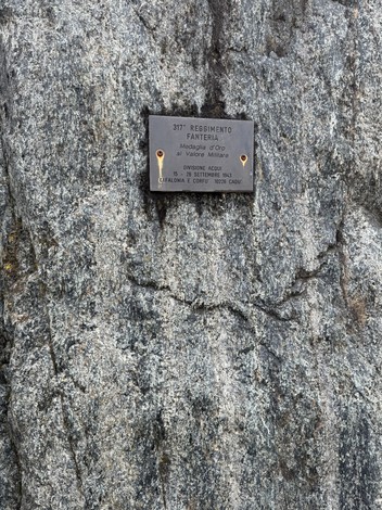 Targa metallica con iscrizione fissata su una parete di roccia grigia.