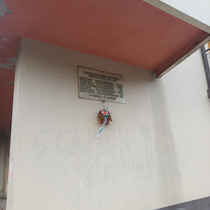 Targa con iscrizione sotto una tettoia, sotto un mazzo di fiori appeso alla parete esterna.