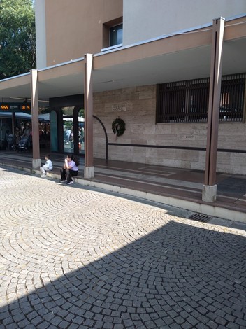 Lapide commemorativa su una parete di pietra con persone sedute in un'area pubblica.