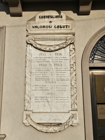 Lapide commemorativa in marmo dedicata ai caduti, adornata da dettagli decorativi e incisa con nomi.