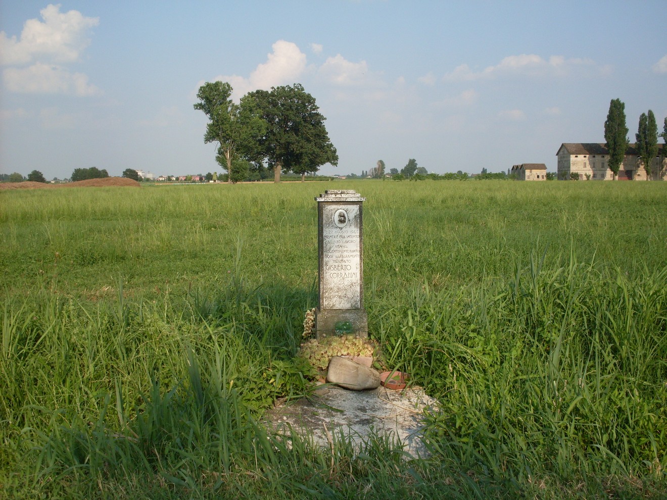 Cippo commemorativo in un prato verde, circondato da alberi e case sullo sfondo.