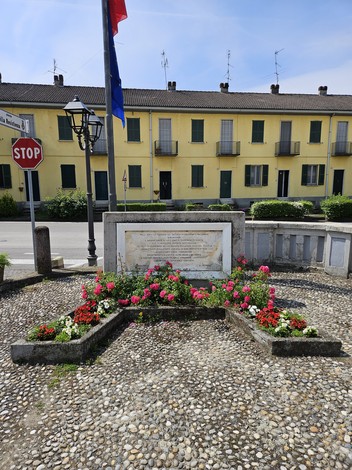 Monumento alla Brigata Balladore, circondato da fiori e cartelli stradali, in un contesto urbano.