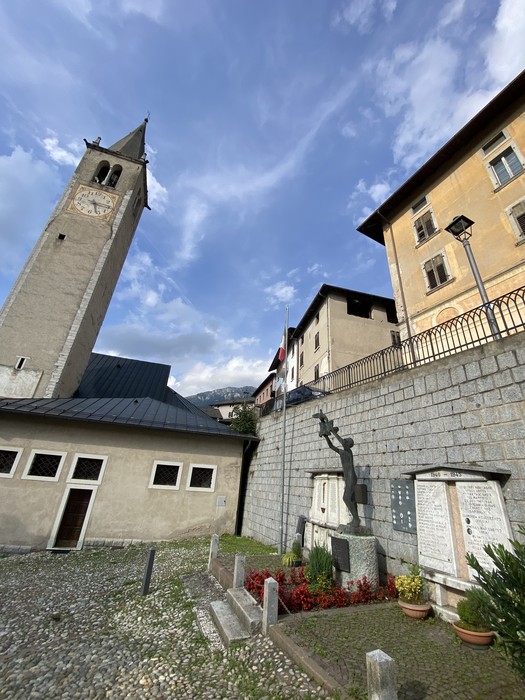 Monumento ai caduti di Castello Tesino, con torre campanaria e muratura in pietra sullo sfondo.
