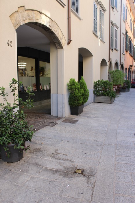 Pietra d'inciampo in un vicolo con piante e un edificio caratteristico sullo sfondo, a Ghezzi.