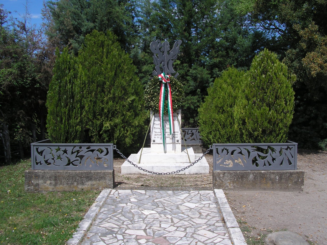 Monumento commemorativo a Ponte del Ricci, circondato da alberi e decorato con un nastro tricolore.