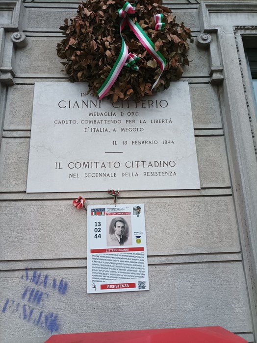Lapide in marmo con iscrizione e corona commemorativa, fissata su una parete in pietra.