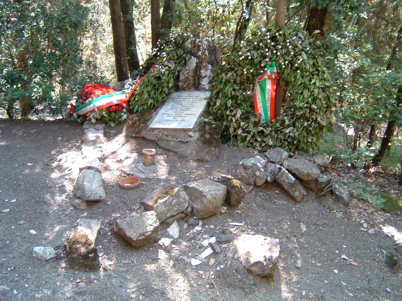 Cippo commemorativo dedicato ai caduti di Radio Cora, circondato da decorazioni e piante nel bosco.