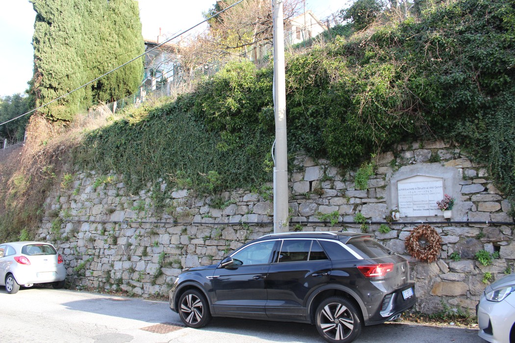 Lapide commemorativa ai caduti di Begato, collocata su un muro di pietra con vegetazione circostante.