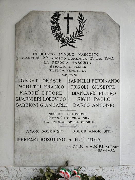 Lapide marmorea a parete con corona d'alloro e croce scolpite sopra una lunga iscrizione, fiori sotto.