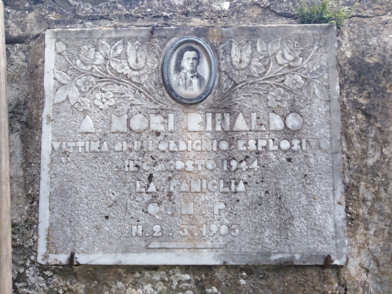 Targa commemorativa con scritte e ritratto, dedicata a Mori Rinaldo, situata in un contesto naturale.